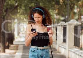 One young mixed race woman standing in the city and using her cellphone to listen to music through headphones while drinking a takeaway coffee. Hispanic woman browsing the internet on a phone downtown