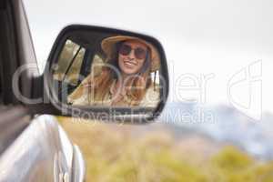 Reflection in the rear view mirror of a young beautiful womans car while travelling to a vacation on a sunny day. Caucasian woman wearing sunglasses and a hat inside a car going on a roadtrip looking at her reflection and smiling while driving on the road