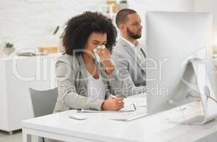 Young mixed race businesswoman blowing her nose with a tissue while sitting at a desk at work. Sick hispanic female businessperson with a curly afro writing on paper while suffering from allergies at work