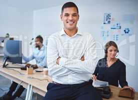 Portrait of confident young mixed race call centre telemarketing agent standing with arms crossed while working in a call centre with colleagues in the background. Happy male manager and supervisor operating helpdesk for customer service and sales support