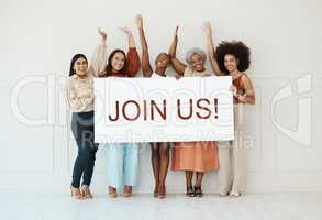 Group of five diverse young businesswomen standing against a wall in an office and holding a recruit sign. Happy colleagues holding a sign with a message while standing in a row together at work