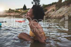 Cheerful African american woman with afro wearing a bikini while sitting in lake. Carefree woman laughing and having fun while out for a swim in river, lake or beach