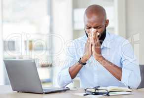 Young african american businessman blowing his nose with a tissue while sitting at a desk at work. Sick male businessperson using a laptop while suffering from allergies at work