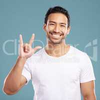 Handsome young mixed race man gesturing rock on while standing in studio isolated against a blue background. Hispanic male showing the sign language of I love you to show affection or romance
