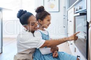 Happy young loving mother and her daughter baking together and having fun while sitting by the oven and watching their homemade cake or cookies bake