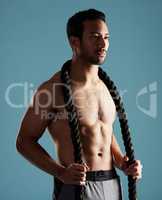 Handsome young hispanic man training with battle ropes in studio isolated against a blue background. Mixed race shirtless male athlete exercising or working out to increase his strength and fitness
