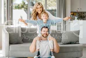 Portrait of playful and excited little boy sitting on his fathers shoulders and holding mothers hands while pretending to fly for fun at home. Happy caucasian parents bonding and relaxing with son