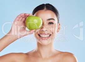 Beautiful young mixed race woman with an apple isolated in studio against a blue background. Her skincare regime keeps her fresh. For glowing skin, eat healthy. Packed with vitamins and nutrients