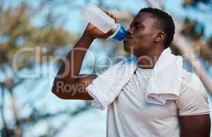 Young active black male athlete holding a bottle and drinking water with a towel around his neck in a forest outside. Runny feeling dehydrated quenching his thirst with some water on sunny hot summers day