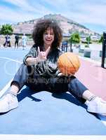 Trendy young hispanic woman making the rock gesture with her hands and fingers, holding a basketball on the court. Happy young woman with afro sitting on the basketball court and showing hand gesture