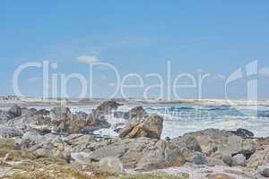 A shallow rocky coastline in the Cape Province, South Africa. Ocean waves crashing on coastal rocks and boulders on a sunny summer day, blue clear skies, and a scenic tropical landscape beachfront