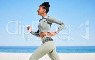 Fit and active African American woman listening to music through earphones and running alone on a beach for workout. Focused black athlete training, exercising and using a cellphone to track progress