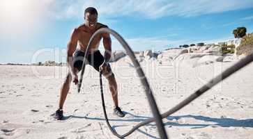 Full length fit and active african american man using battle ropes to workout on a beach alone in the day. Strong and muscular black man training shirtless in nature. Focused on health and strength