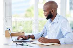 Young african american businessman writing in a notebook and working on a laptop in an office alone at work. One male businessperson using a laptop at a table at work