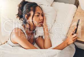 A young mixed race woman looking depressed while scrolling social media and lying in bed. An attractive Hispanic female using her cellphone and looking exhausted while resting in her bedroom