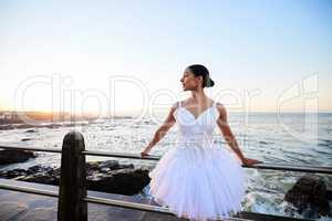 Elegant young ballerina wearing white fancy dress while leaning against a railing on the promenade with a beautiful ocean background at sunset