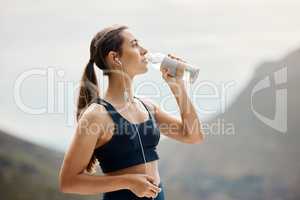 One fit young mixed race woman taking a rest break to drink water from bottle while exercising outdoors. Female athlete wearing earphones quenching thirst and cooling down after running and training workout