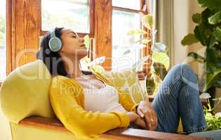 Woman listening to music on headphones while comfortable and relaxing in a bright living room. A young hispanic female with eyes closed resting and sitting on a chair using modern technology at home