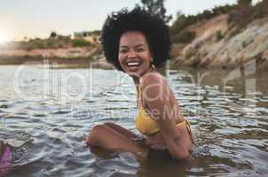 Portrait of cheerful African american woman with afro wearing a bikini while sitting in water outdoors. Carefree woman having fun while out for a swim at the lake
