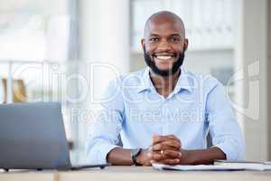 Portrait of a young happy african american businessman sitting at a desk in an office alone at work. One male businessperson smiling while using a laptop at a table at work