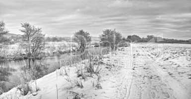 A black and white view of a mountain lake in winter, covered with snow and ice. The beautiful cloudy weather around the pond covered with snowfall. A dense forest and cloudy sky on a glacial valley.