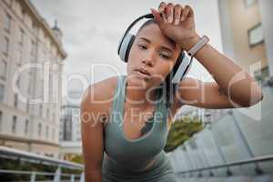 Portrait of a young mixed race female athlete wearing wireless headphones and listening to music while standing outside in the city wiping her sweaty forehead and tired from a workout. Determined to get a fit and toned body