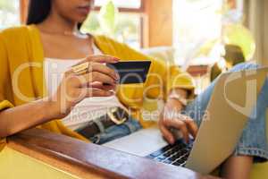 Unrecognizable woman shopping online while typing on her laptop and holding her bank card sitting on a chair in a bright living room. A hispanic young female at home using modern technology for buying