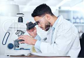 One serious young male medical scientist sitting at a desk and using a microscope to examine and analyse test samples on slides in a hospital. Hispanic healthcare biochemist professional discovering and innovating a cure for diseases in his laboratory