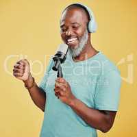 Happy mature African American man against a yellow studio background wearing headphones to listen to music while singing karaoke with a microphone. Smiling black man enjoying and holding a mic to sing