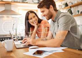 Young cheerful interracial couple bonding while working on a laptop together at home. Caucasian boyfriend and girlfriend working through their bills and using a laptop. Content wife staring at her husband while typing on a computer
