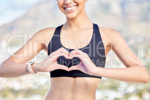 Closeup of one confident young mixed race woman gesturing a heart shape with her hands while exercising outdoors. Happy female athlete caring for body with regular training workout or run. Endorsing a healthy active lifestyle