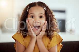 Adorable little mixed race child touching face with her hands in surprise at home. One small cute hispanic girl sitting alone on a living room sofa and looking shocked. Excited kid with curly hair