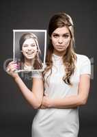 Different day, different mood. Studio shot of an attractive young woman dressed up in 60s wear and holding a photograph of herself smiling against a dark background.