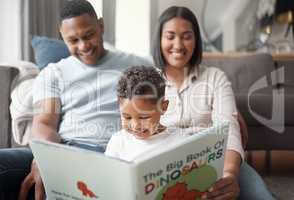 A happy mixed race family of three relaxing on the lounge floor and reading a story book at home. Loving black family being affectionate on a carpet. Young couple bonding with their son at home