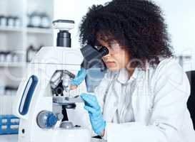 Young african american woman wearing a labcoat and goggles looking at medical samples on a microscope in her lab. A mixed race female scientist wearing goggles and gloves conducting research