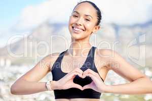 Portrait of one confident young mixed race woman gesturing a heart shape with her hands while exercising outdoors. Happy female athlete caring for body with regular training workout or run. Endorsing a healthy active lifestyle