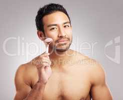 Handsome young mixed race man posing shirtless in studio isolated against a grey background. Hispanic male using a dermal face roller to massage and relax his skin. All part of his skincare regime