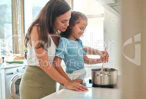 Adorable little girl and her mother cooking together at home. Young mother standing behind her daughter and helping her while stirring food on the stove. Mom and child preparing dinner together