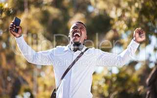 African American man celebrating a win while using his cellphone and wearing headphones. Happy young male expressing success and victory while walking outside. Good news makes this man smile