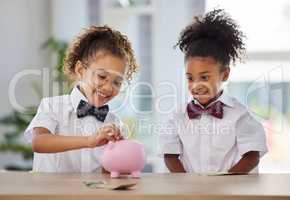 Savings go a long way. two adorable little girls dressed as businesspeople sitting in an office and depositing money into a piggybank.