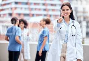 Portrait of a happy young doctor gesturing thumbs up with her colleagues in the background. Group of doctors and nurses collaborating in meeting. Powerful medical professional giving thumbs up