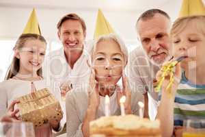 Senior woman celebrating a birthday with family at home, wearing party hats and blowing whistles. Grandma looking at birthday cake and being joyful while surrounded by her grandkids, husband and son