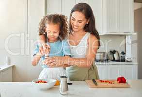 Happy mother teaching little daughter to cook in kitchen at home. Little girl adding seasoning pepper grinder to a bowl while making a salad with mom