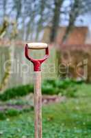 Shovel in a green garden outdoors in a backyard of a house. Closeup of a wooden gardening tool or equipment outside in a yard with a fence and trees in the background. A spade stuck in the ground