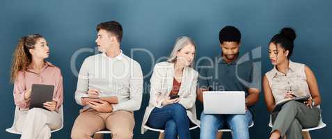 Group of diverse serious businesspeople sitting on chairs in a row in an office together. Five business professionals sitting and talking using technology in a waiting room together in an office