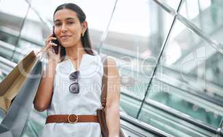 Trendy young woman talking on a cellphone during a shopping spree in a mall. One female only enjoying retail therapy while connecting on a smartphone. Happy shopper holding bags and making a call on a escalator