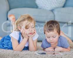 Young caucasian boy and girl watching cartoon videos online and playing fun games on digital tablet to learn from educational apps while lying on the carpet floor at home. Two kids using smart gadget