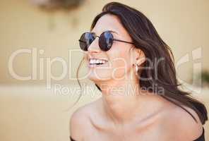 Close up of cheerful mixed race woman wearing sunglasses and laughing while standing outside on sidewalk. Joyful young brunette woman looking stylish in a off-shoulder top on a summer day