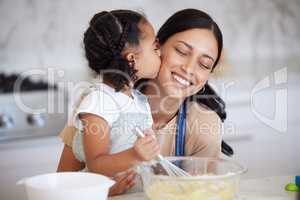 Happy latin mother and daughter baking and sharing a kiss while bonding. A young woman helping her daughter bake or cook while stirring the batter and giving an affectionate hug in the kitchen