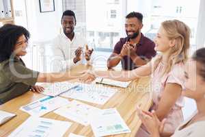 Female coworkers shaking hands while their colleagues clap hands. Group of diverse businesspeople having a meeting at a table in an office. Business professionals talking and planning together at work